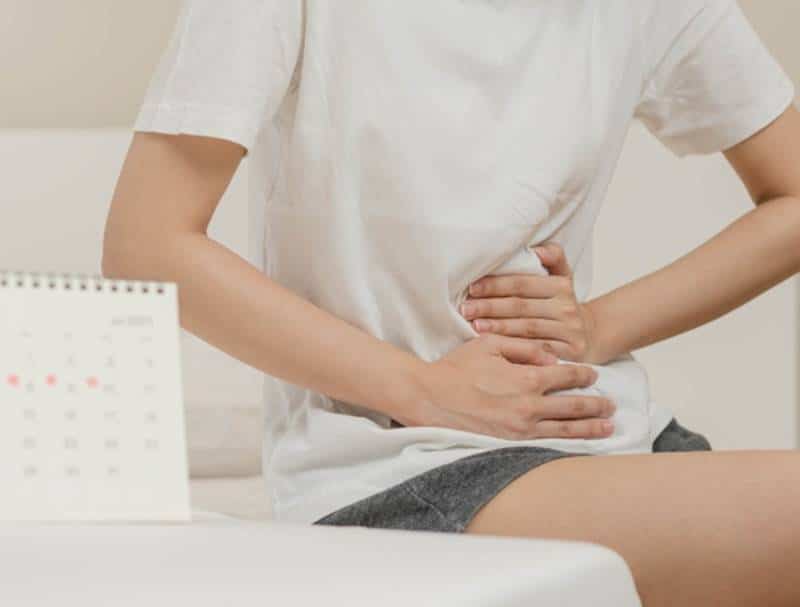 의자에 앉아 아랫배를 손으로 누르고 있는 모습으로, 생리 전 체내 수분 정체와 함께 나타나는 복부 붓기 증상을 설명하는 이미지.