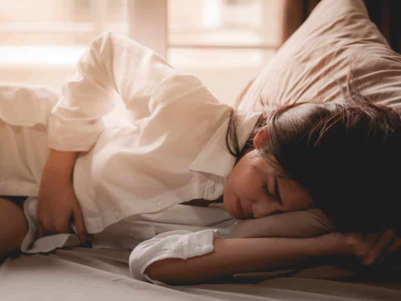 생리 전 컨디션 저하로 복부가 불편해 보이는 상태에서 옆으로 누워 휴식을 취하는 모습으로, 생리 전 붓기와 몸의 변화 상황을 표현한 이미지.