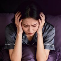 잠을 충분히 자지 못해 침대에서 피곤한 표정으로 머리를 감싸고 앉아 있는 여성의 모습으로, 수면 부족으로 인해 얼굴 붓기가 생길 수 있는 상황을 표현한 이미지.