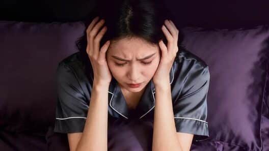 잠을 충분히 자지 못해 침대에서 피곤한 표정으로 머리를 감싸고 앉아 있는 여성의 모습으로, 수면 부족으로 인해 얼굴 붓기가 생길 수 있는 상황을 표현한 이미지.