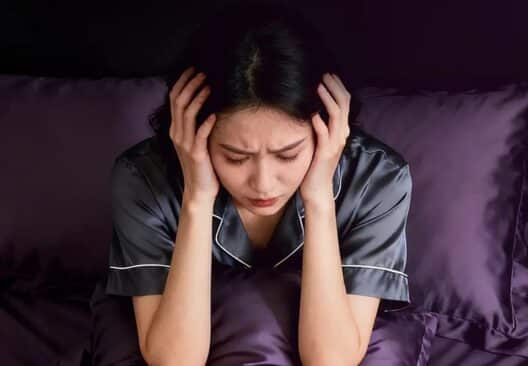 잠을 충분히 자지 못해 침대에서 피곤한 표정으로 머리를 감싸고 앉아 있는 여성의 모습으로, 수면 부족으로 인해 얼굴 붓기가 생길 수 있는 상황을 표현한 이미지.