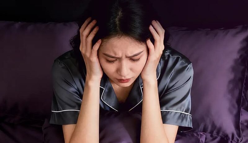 잠을 충분히 자지 못해 침대에서 피곤한 표정으로 머리를 감싸고 앉아 있는 여성의 모습으로, 수면 부족으로 인해 얼굴 붓기가 생길 수 있는 상황을 표현한 이미지.