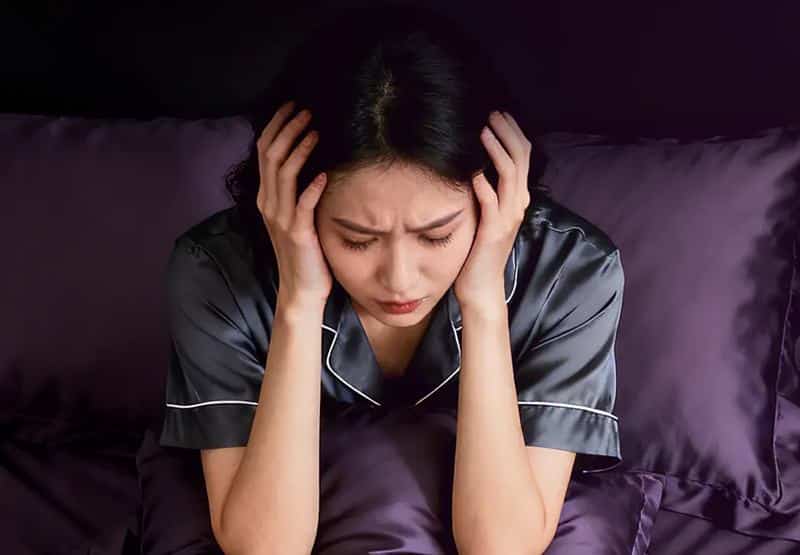 잠을 충분히 자지 못해 침대에서 피곤한 표정으로 머리를 감싸고 앉아 있는 여성의 모습으로, 수면 부족으로 인해 얼굴 붓기가 생길 수 있는 상황을 표현한 이미지.